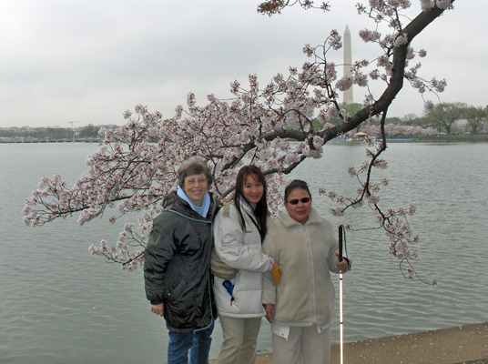 Photo shows Dona, Nanta and Ann in front of water with a branch of cherry blossons hanging over and behind them.  On the other side of the water is land with short buildings, behind which the tall, slender Washington Monument can be seen.  Ann is holding Nanta's arm and Nanta is holding a white cane.